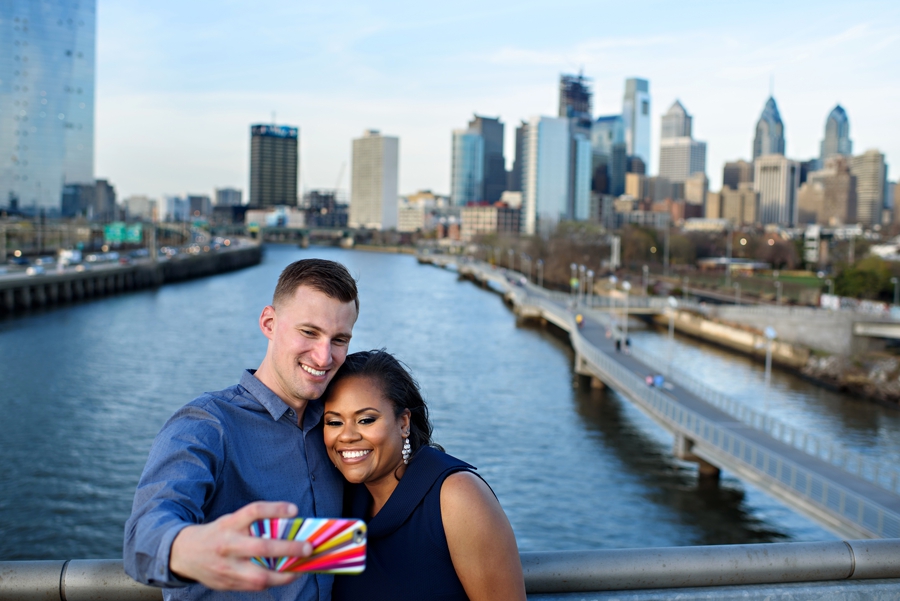 An engaged couple take a selfie on south street bride with the Philly skyline behind them.